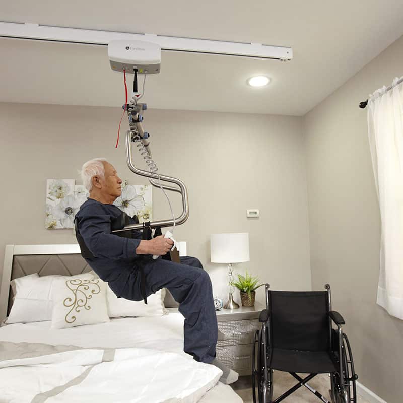 Elderly person using a ceiling-mounted Handicare C-450 ceiling lift system in a bedroom with a bed and wheelchair.