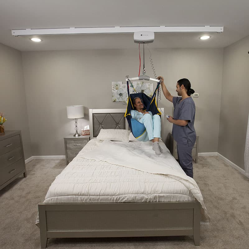 Person in a blue sling being assisted by a caregiver in a bedroom with a ceiling hoist system.