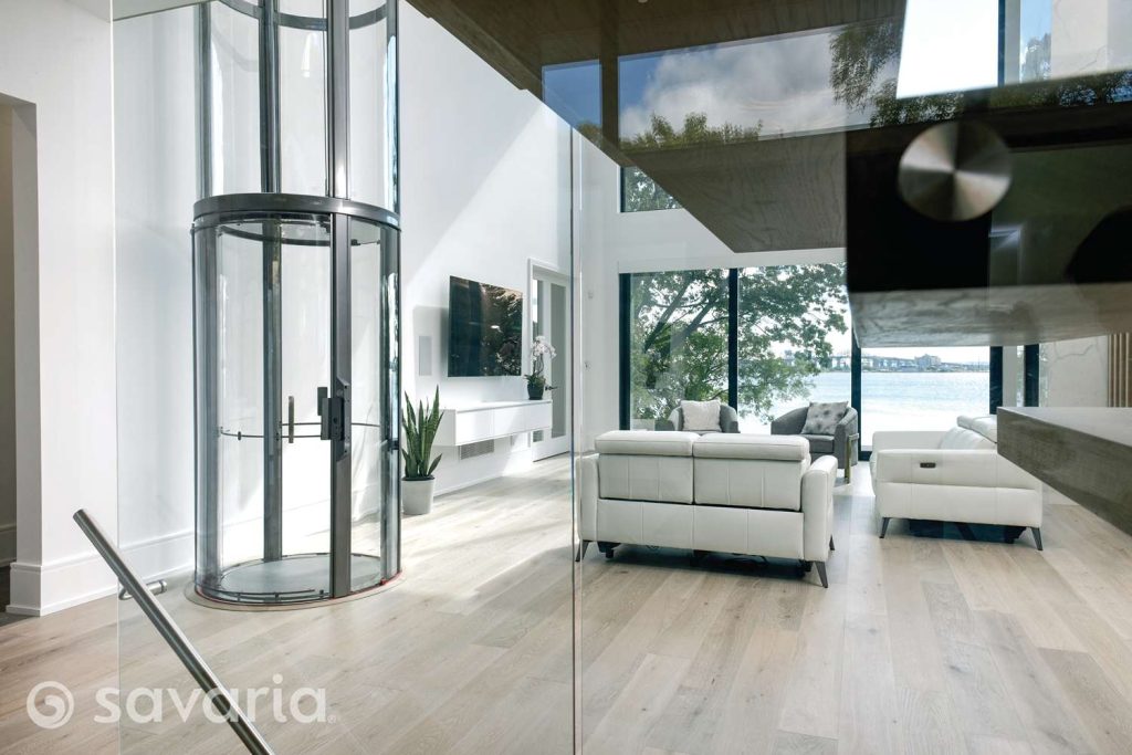 Modern living room with a glass elevator and large windows overlooking water.